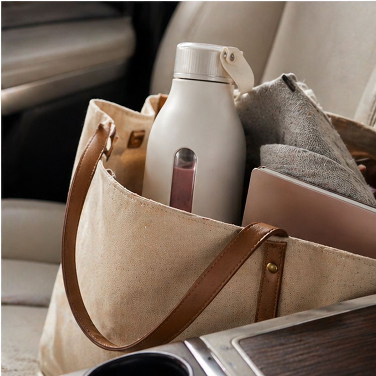 Beige tote bag with brown leather straps containing a white water bottle, gray towel, and pink item on a car seat.